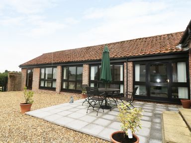 A patio area with a round table, chairs, and a closed umbrella outside a brick building with tiled roof at Jacko's Barn in Homersfield