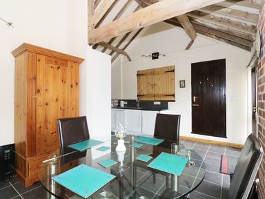 A dining area with a glass table and blue placemats near a wooden cupboard and a kitchen with white cabinets at Jacko's Barn in Homersfield