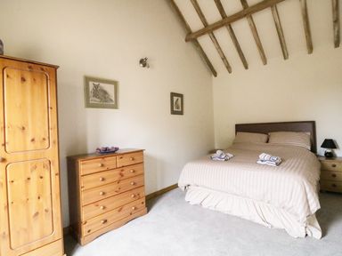 A bedroom with a double bed, wooden wardrobe, chest of drawers, bedside table with lamp and framed pictures on the wall at Jackos Barn in Homersfield