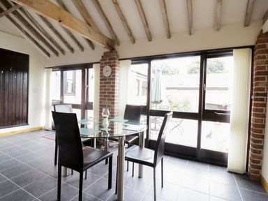 A dining area with a glass table and four black chairs near large windows with vertical blinds at Jacko's Barn in Homersfield