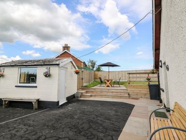 An outdoor area with a small building a wooden bench flower pots a table with chairs and an umbrella at Green Cottage in Pant-y-Dwr near Rhayader