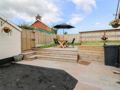 A backyard patio with a wooden table, chairs, an umbrella, and hanging flower baskets at Green Cottage in Pant-y-Dwr near Rhayader