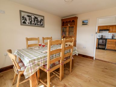 A dining room with a wooden table covered with a checkered tablecloth and six chairs near a wooden cabinet and a kitchen visible through the doorway at Green Cottage in Pant-y-Dwr near Rhayader