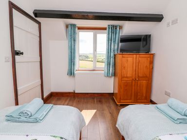 A bedroom with two single beds with folded towels on them a wooden wardrobe with a television on top a window with curtains and a door at Green Cottage Pant-y-Dwr near Rhayader