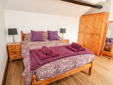 A bedroom with a double bed with purple bedding and towels two bedside tables with lamps a wooden wardrobe and a standing mirror at Green Cottage in Pant y Dwr near Rhayader