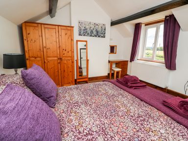 A bedroom with a patterned bedspread purple cushions wooden wardrobe and a dressing table with curtains at Green Cottage Pant-y-Dwr near Rhayader