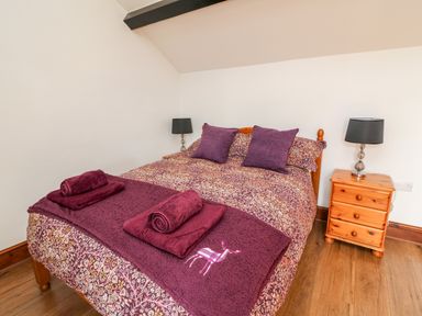 A bedroom with a bed covered with patterned bedding and purple cushions and towels next to wooden bedside tables with lamps at Green Cottage Pant-y-Dwr near Rhayader