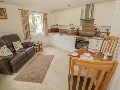 A kitchen and living area with a brown leather sofa wooden dining table set with dishes and glassware and white cabinets at Lawn Lodge in Ashleworth