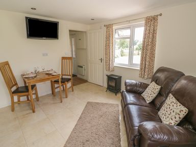 A small living room with a brown leather sofa and two cushions a wooden dining table with two chairs a wall-mounted tv and a window with patterned curtains at Lawn Lodge in Ashleworth