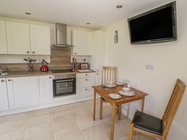 A kitchen with white cabinets a wooden dining table set for two and a wall-mounted TV at Lawn Lodge in Ashleworth