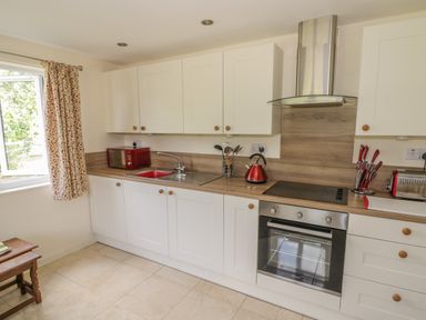 A kitchen with white cabinets a wooden countertop a red microwave red kettle red toaster and a window with patterned curtains at Lawn Lodge in Ashleworth