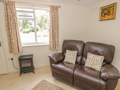 A room with a brown leather sofa with two patterned cushions a window with patterned curtains an electric stove heater and a framed fruit painting on the wall at Lawn Lodge in Ashleworth