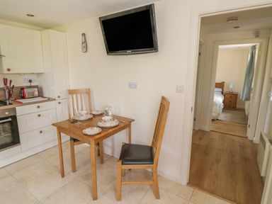 A kitchen with a small wooden dining table set for three and a wall-mounted TV with a view into a bedroom with a wooden bed and nightstand at Lawn Lodge in Ashleworth