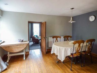 A dining room with a round table covered with a cloth and four chairs and a wooden side table near an open door at Corner Cottage in Reynoldston near Swansea