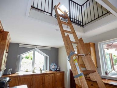 A wooden ladder leading to a loft opening in a kitchen with wooden cabinets and windows at Corner Cottage in Reynoldston near Swansea