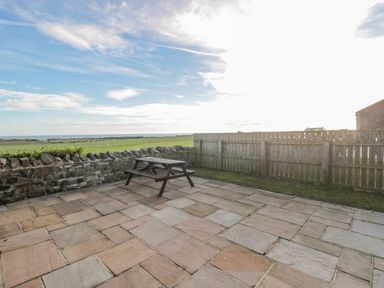 A patio with a wooden picnic table and stone wall overlooking a grassy field and sea at Long Cart Cottage in Embleton