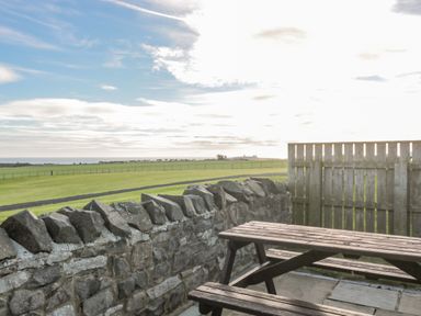 An outdoor wooden picnic table and benches on a stone patio with a stone wall and a wooden fence overlooking grassy fields