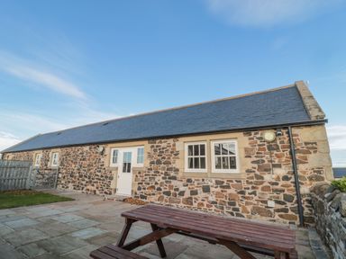 An outdoor patio area with a wooden picnic table in front of a stone building with a white door and windows at Long Cart Cottage in Embleton