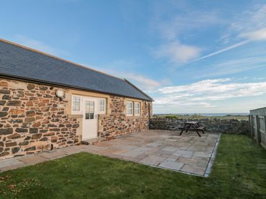 Exterior of a stone building with a white door and windows patio with picnic table lawn and stone wall at Long Cart Cottage in Embleton