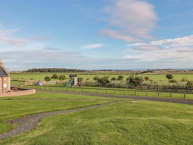 A grassy area with a children's play structure and a small building on the left with fields and trees in the background at Long Cart Cottage in Embleton