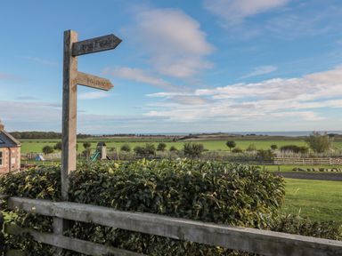 A wooden signpost with directions to Embleton and to beach in a rural setting with fields and playground equipment at Long Cart Cottage in Embleton