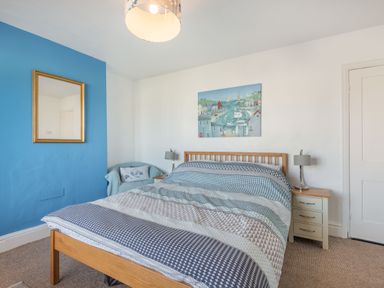 A bedroom with a wooden bed frame and patterned bedding a blue accent wall with a mirror an armchair a bedside table with a lamp and a painting above the bed at 2 Tregof Terrace in Cemaes Bay