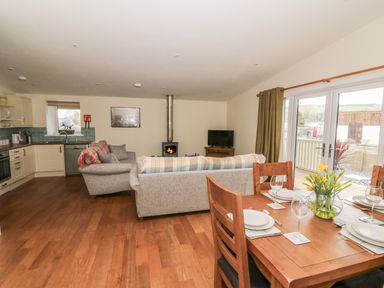 An open plan kitchen and living room with sofas a wood burner a dining table set for four and glass doors leading outside at Y Felin - Moana in Borth near Aberystwyth