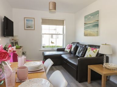 A living room with a brown leather sofa floral cushions a wooden side table with a lamp and a dining table set with plates cups and pink flowers at White Rose Apartment in Filey