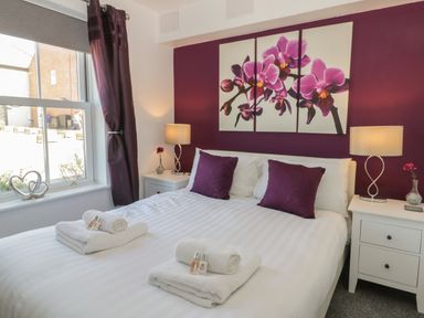 A bedroom with a double bed two bedside tables and lamps purple cushions and floral wall art at White Rose Apartment in Filey