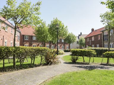 A paved pathway through a garden with trimmed hedges and trees between brick houses at White Rose Apartment in Filey