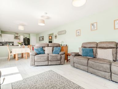 A living room with two grey sofas blue and grey cushions a dining table with chairs and a kitchen area at Kingfisher Lodge in Brandesburton