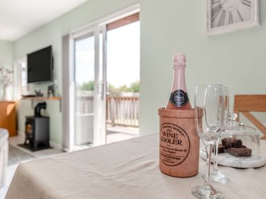 A dining table with a wine cooler holding a bottle and two glasses in a room with a wood stove and a wall mounted tv at Kingfisher Lodge in Brandesburton