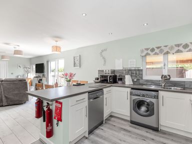A kitchen with white cabinets a dishwasher a washing machine a grey countertop and a dining area with chairs and flowers at Kingfisher Lodge in Brandesburton