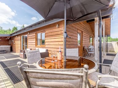 An outdoor wooden deck with a round glass table, chairs, a large umbrella, and a wooden cabin wall at Kingfisher Lodge in Brandesburton