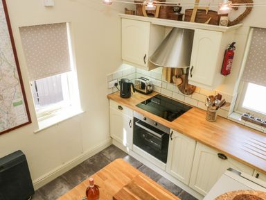 A kitchen with white cabinets wooden countertops a black electric stove a toaster kettle utensil holder two windows with dotted blinds and a fire extinguisher at Gable End Coniston near Hawkshead