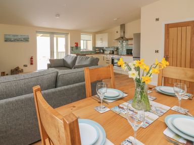 A dining table set with plates and glasses with a vase of flowers and a kitchen with grey sofas in the background at Y Felin - Celyn in Borth