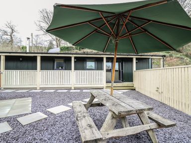 An outdoor patio area with a wooden picnic table and large green umbrella in front of a building with dark green siding and light wood fencing at Y Felin - Celyn in Borth