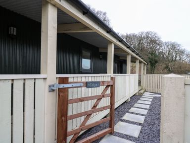 A wooden gate with a sign that reads Celyn leading to a covered porch with a set of concrete stepping stones at Y Felin - Celyn in Borth