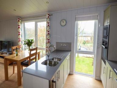 A kitchen and dining area with a sink, countertop, table with chairs, television and windows showing a garden at Crook Lodge in Allithwaite near Grange-over-Sands