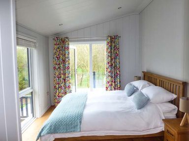 A bedroom with a wooden bed with white bedding and blue pillows next to a window and glass door with colorful curtains at Crook Lodge in Allithwaite near Grange-over-Sands