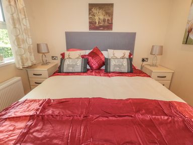 A bedroom with a bed with grey headboard red bedding and decorative pillows and bedside tables with lamps at Little Gem Lodge in Charlesfield near St Boswells
