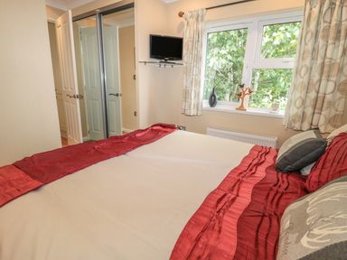 A bedroom with a bed covered with red and white bedding a window with curtains and decorations and a wall-mounted TV at Little Gem Lodge in Charlesfield near St Boswells