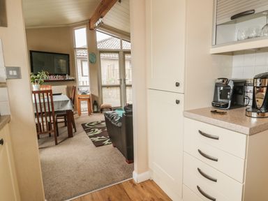 A kitchen area with coffee machines on the counter and white cabinets next to a dining space with a table, chairs, and a television at Little Gem Lodge in Charlesfield near St Boswells