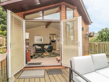 A lounge with sofas and a coffee table seen through open double doors onto a wooden deck with seating at Little Gem Lodge in Charlesfield near St Boswells
