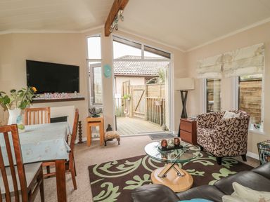 A living room with a dining table chairs a glass coffee table sofa patterned armchair and sliding door to a fenced deck at Little Gem Lodge in Charlesfield near St Boswells
