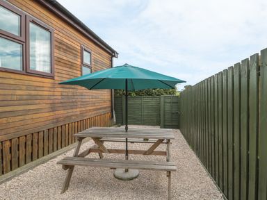An outdoor area with a wooden picnic table and green umbrella next to a wooden building and green fence at Little Gem Lodge in Charlesfield near St Boswells