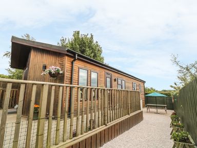 An exterior view of a wooden cabin with multiple windows a wooden railing gravel pathway and a picnic table with a green umbrella Little Gem Lodge Charlesfield near St Boswells