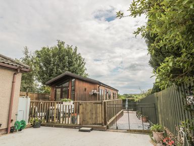A wooden cabin with a fenced patio area and potted plants next to a fenced pathway at Little Gem Lodge in Charlesfield near St Boswells