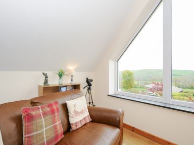 A leather sofa with two cushions next to a window with a countryside view and a small bookshelf with a lamp and decorative items at Treview in Launceston
