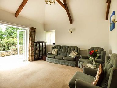 A living room with green upholstered sofas and armchairs a glass cabinet and open patio doors at Shepherds Rest in Jacobstow near Bude